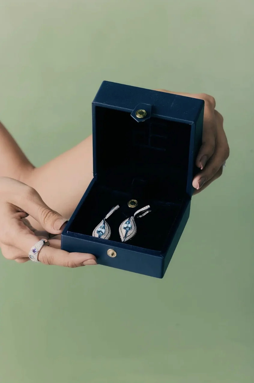 Hands holding an open navy case with a pair of aquamarine and diamond earrings.